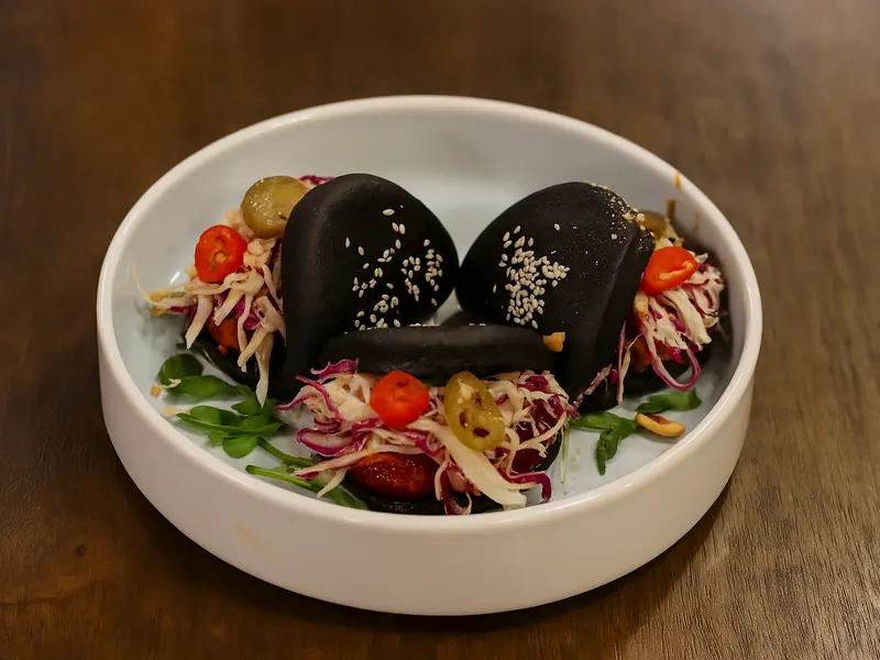Two black sesame-topped bao buns on a white plate, filled with slaw and served with cherry tomatoes and greens.