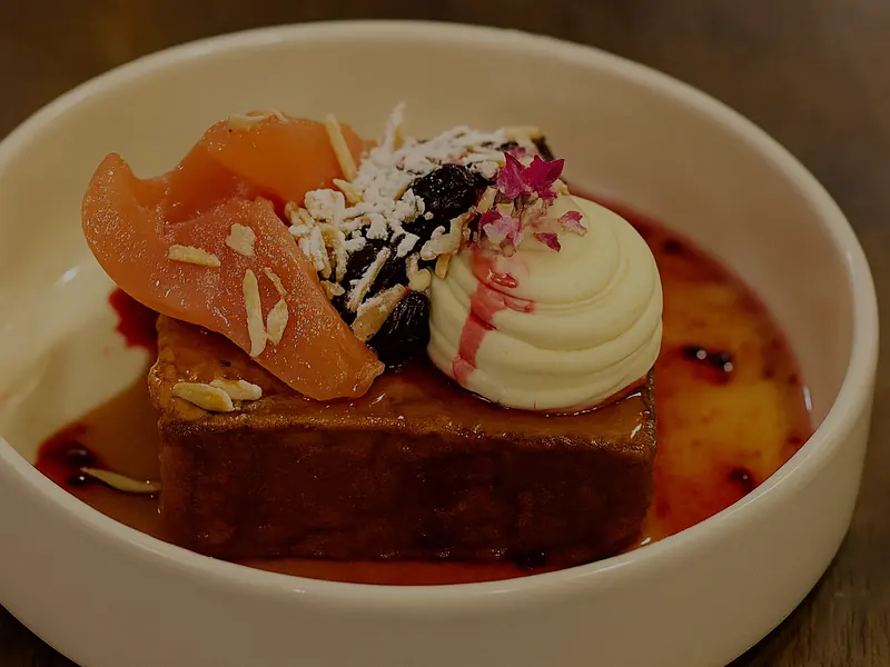 A thick slab of French toast in a white bowl with poached pear slices, a swirl of cream, flaked almonds, coconut and berry syrup.