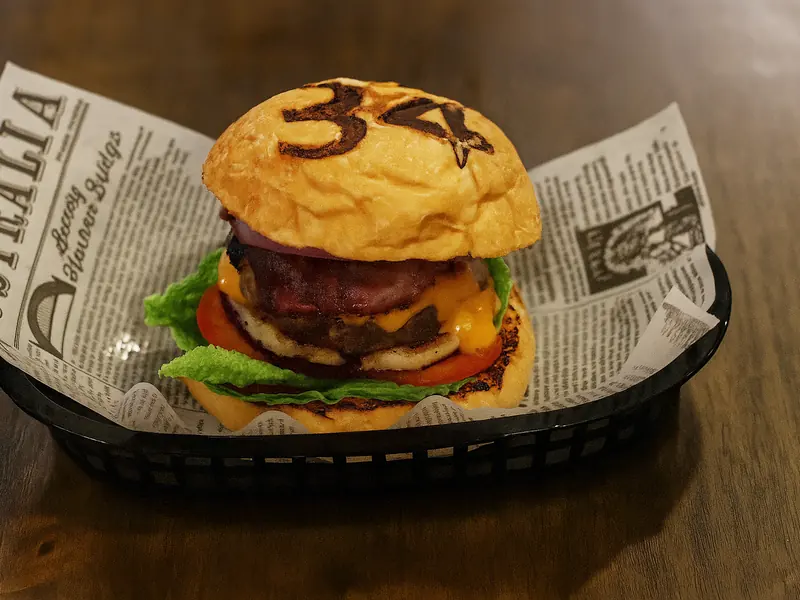 A burger in a 34-branded bun with beef patty, melted cheese, lettuce, tomato and red onion, served in a black basket lined with newsprint paper.